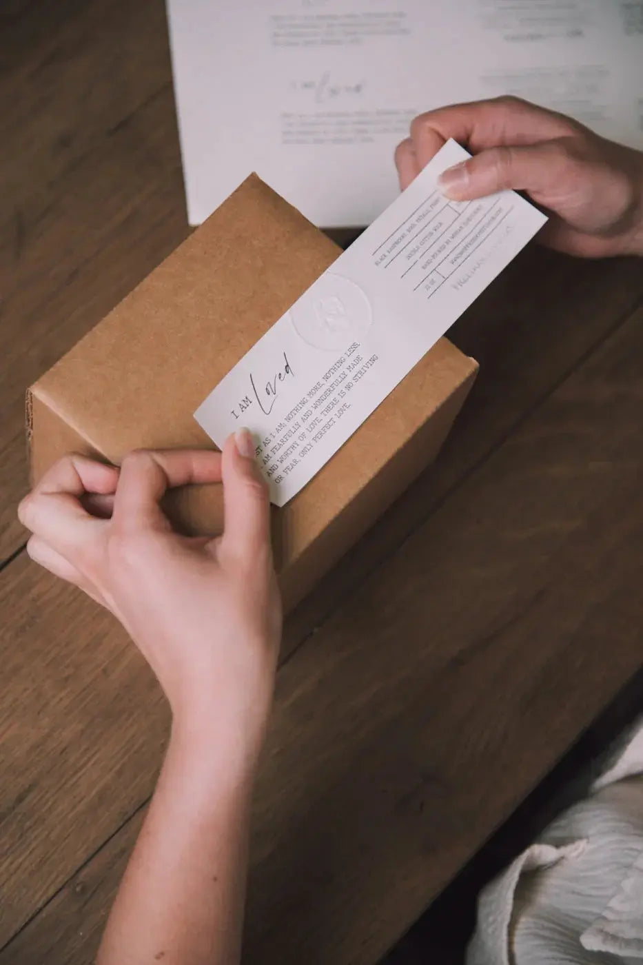 Hands placing a heartfelt 'I Am Loved' note on a rustic kraft gift box, perfect for cozy home decor gifts.