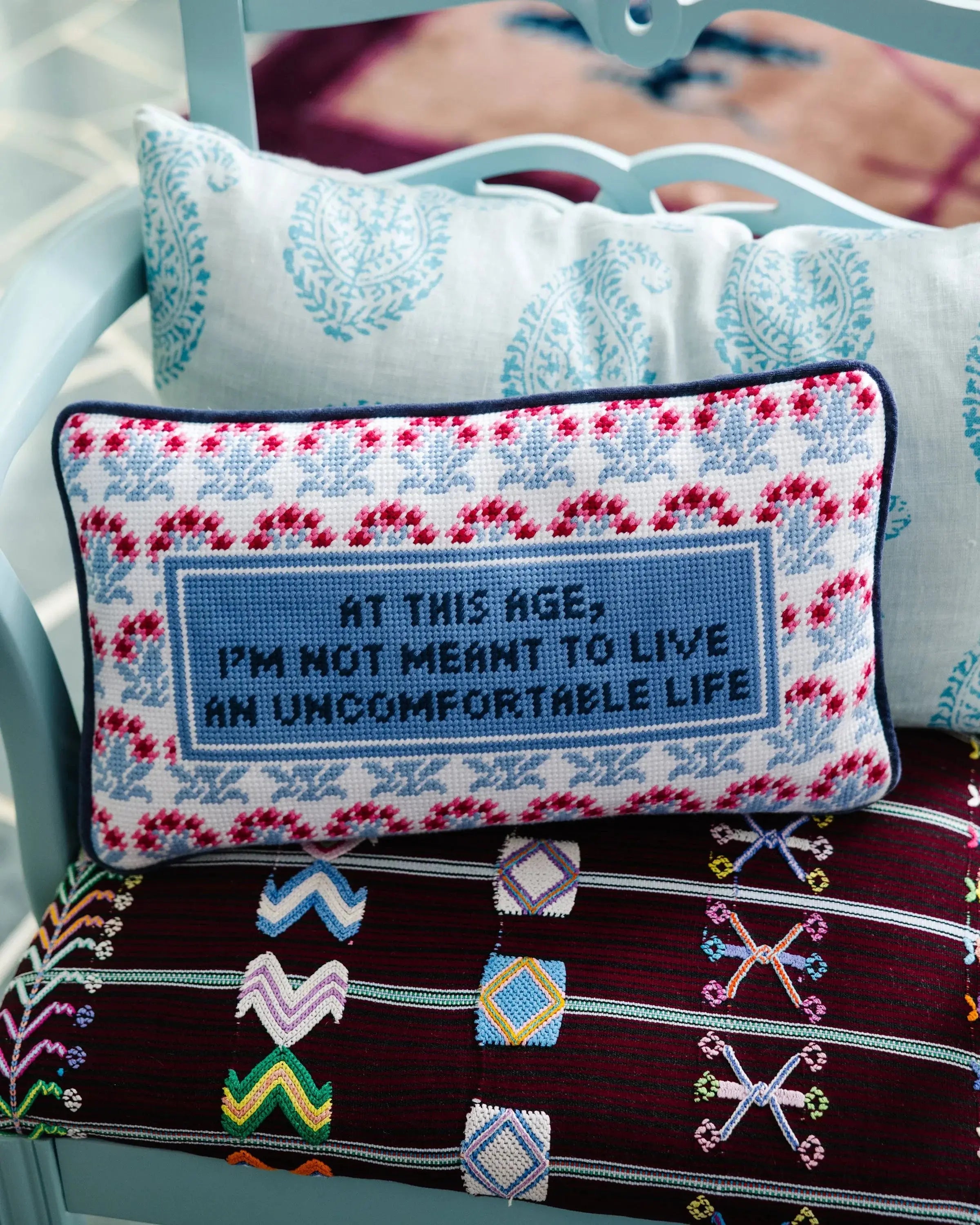 Cozy farmhouse pillow with cross-stitch quote 'At this age, I'm not meant to live an uncomfortable life' on patterned chair