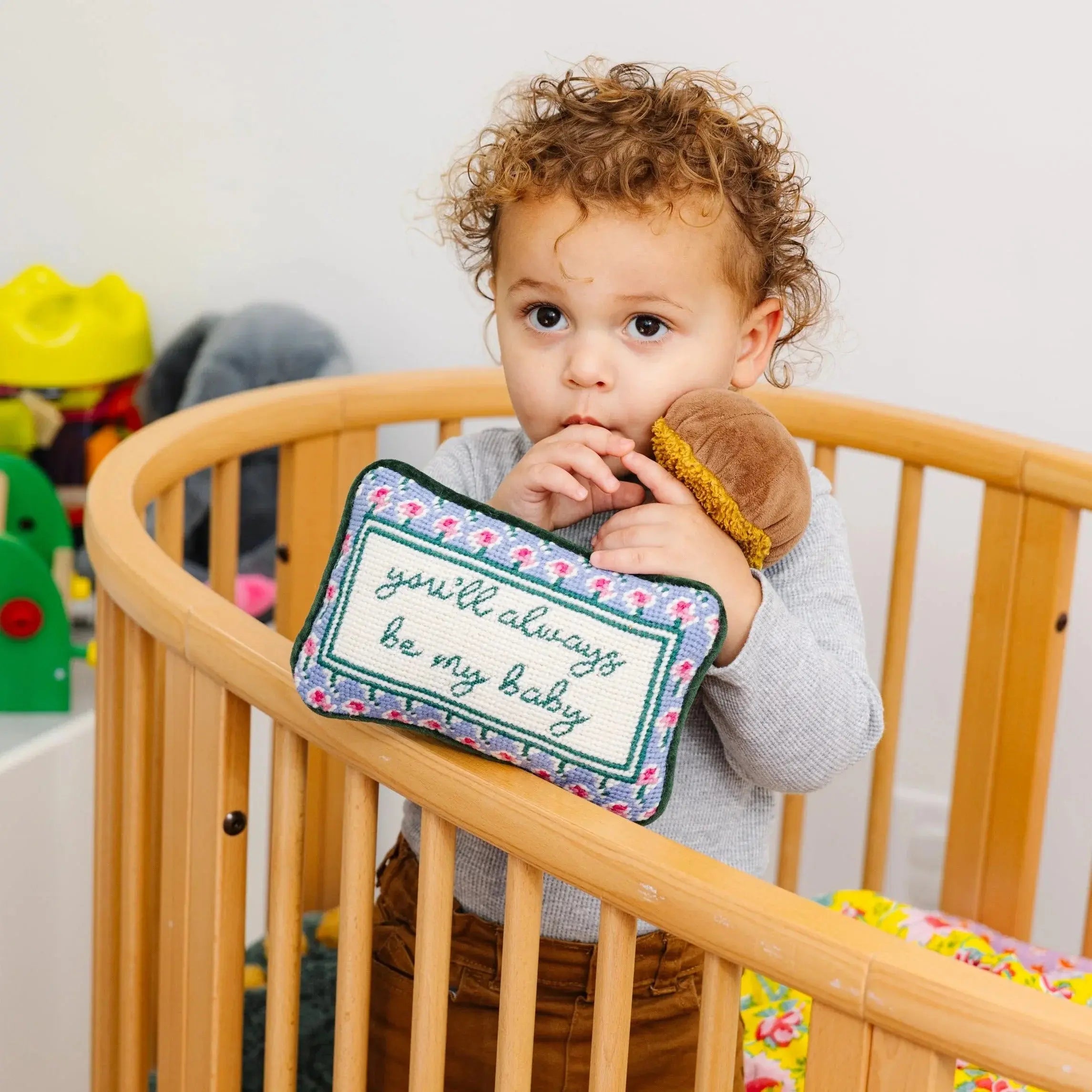 Toddler in wooden crib holding floral needlepoint pillow saying 'you'll always be my baby' — cozy nursery decor
