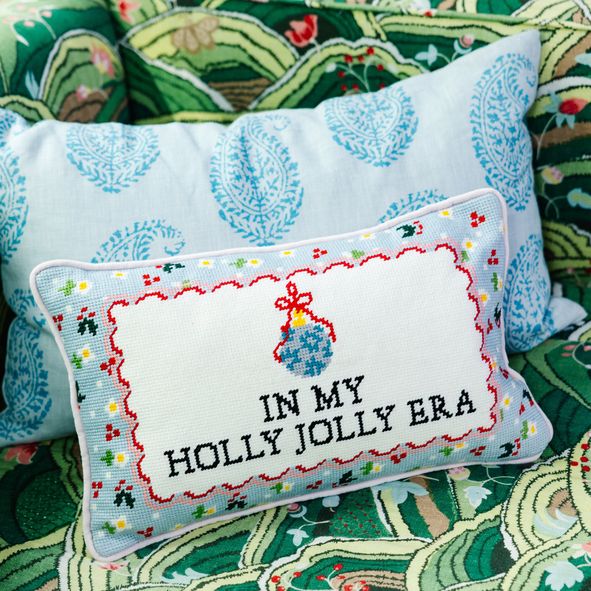A square decorative pillow with a white front panel featuring embroidered text reading 'IN MY HOLLY JOLLY ERA' and a blue hanging ornament motif, styled on top of a larger light blue paisley pillow against a green patterned quilt.