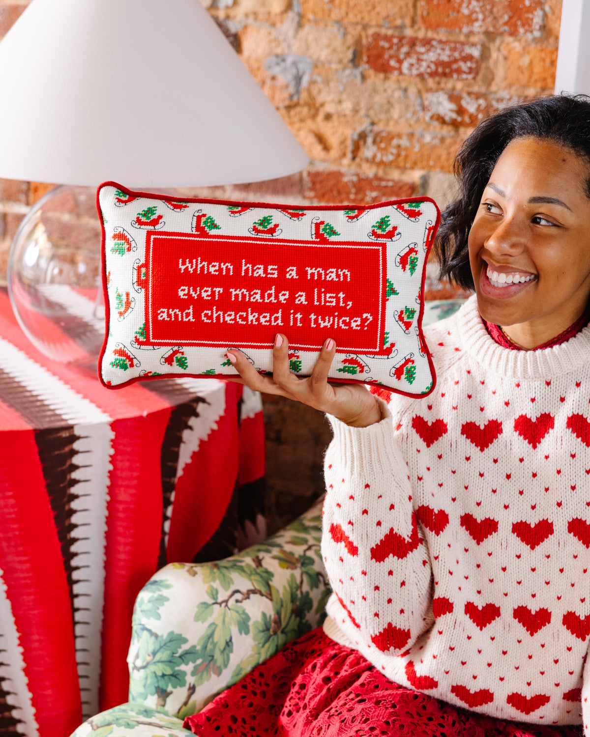 A smiling woman in a red and white heart-patterned knit sweater holds up a rectangular Christmas-themed throw pillow with the text 'When has a man ever made a list, and checked it twice?' on a red background with red chili peppers and green holly accents.