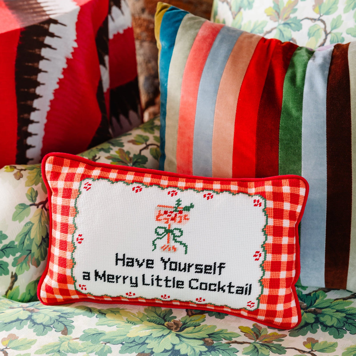 A rectangular decorative throw pillow with a red and white gingham border and a white center panel, embroidered with a jolly green figure and the text 'Have Yourself a Merry Little Cocktail.' It is styled on a floral couch with other colorful pillows in the background.