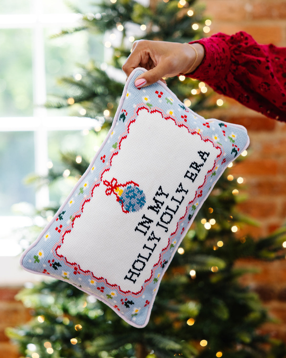 A hand holds a square decorative pillow in front of a blurred Christmas tree and brick wall. The pillow has a light blue border with small embroidered flowers, a blue gift box motif, and the text 'IN MY HOLLY JOLLY ERA' in black on a white center panel.