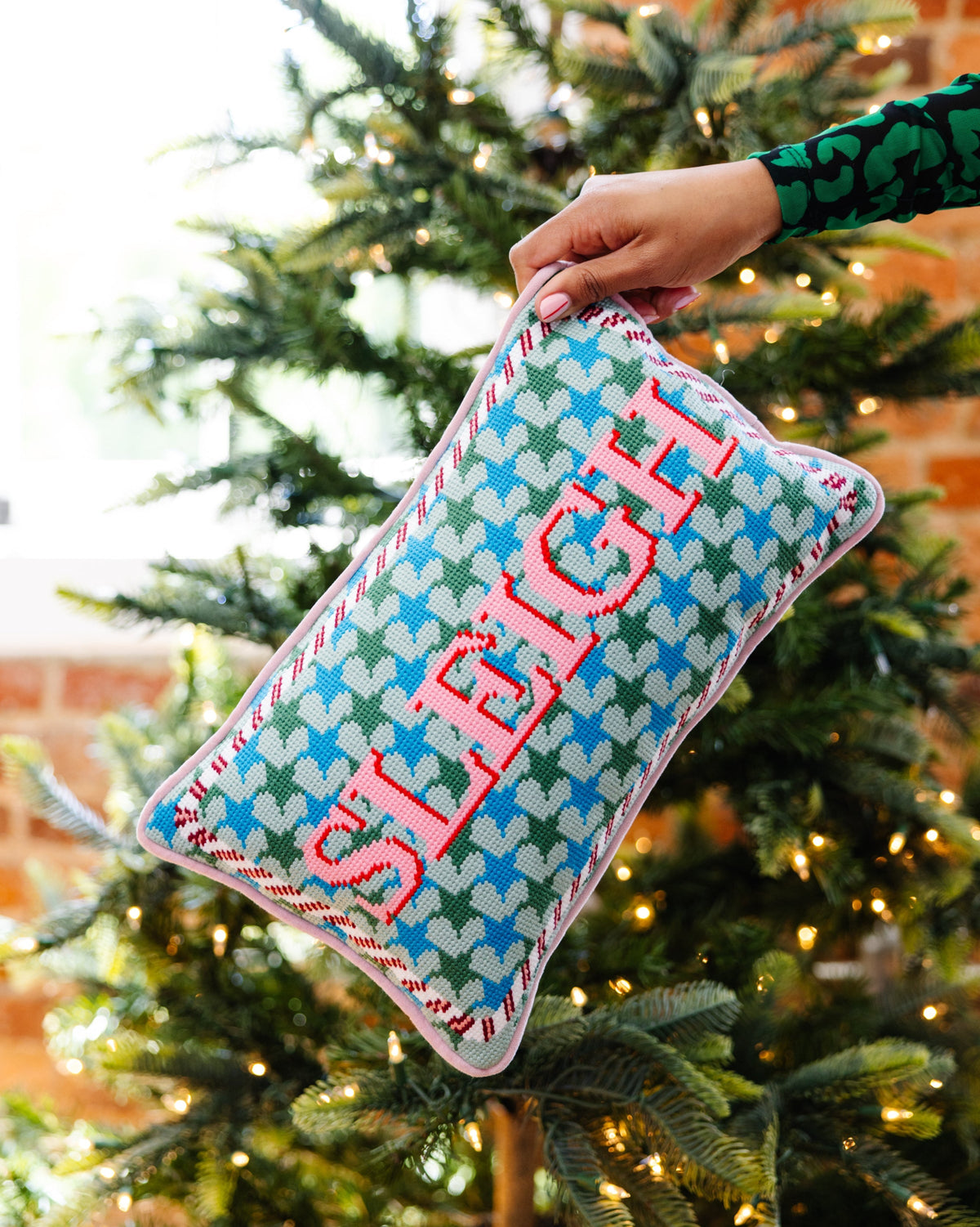 A hand holds a square knitted Christmas pillow with the word 'SLICE' in red letters over a blue, green, and white holly pattern, set against a blurred Christmas tree background.
