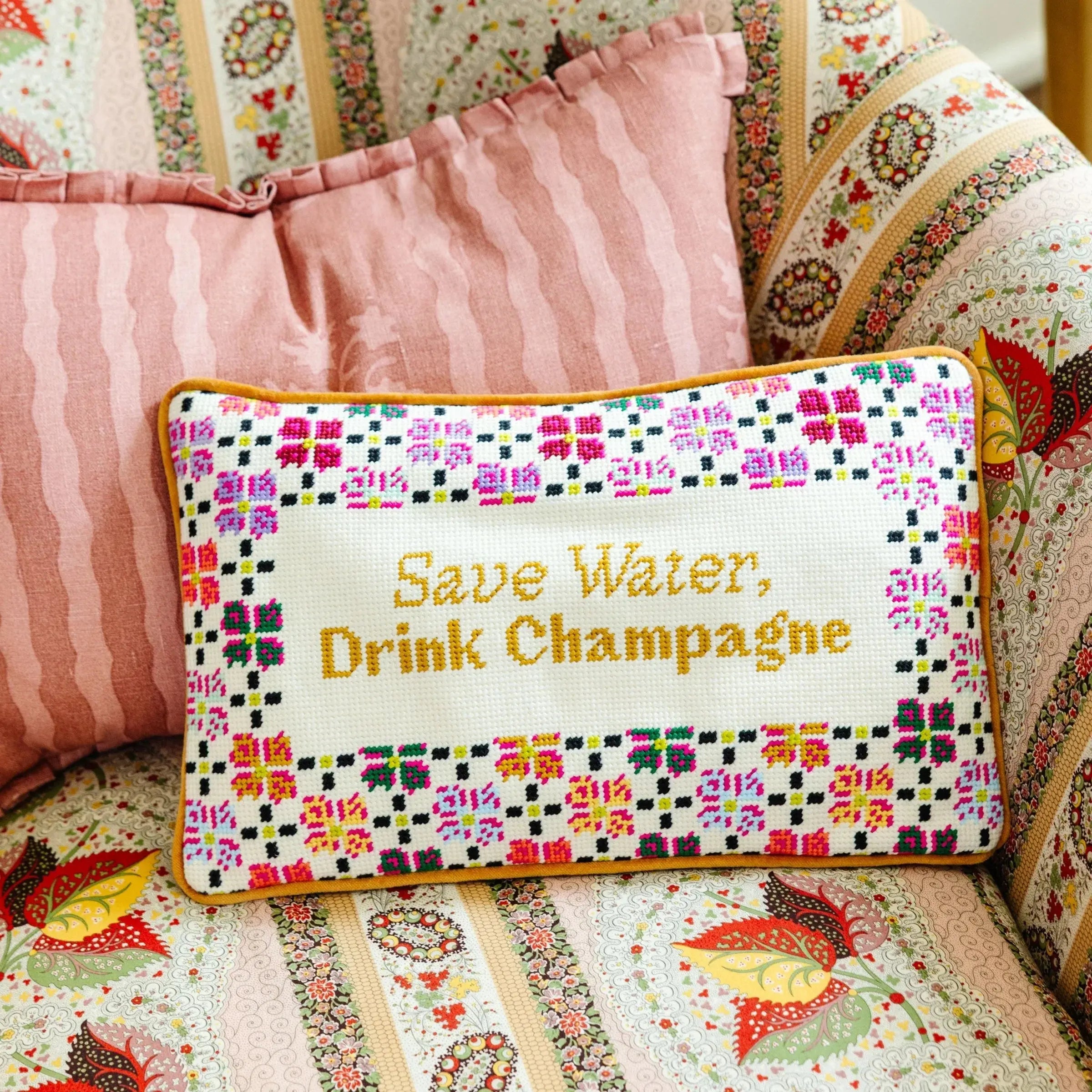 Decorative pillow with colorful pattern and text 'Save Water, Drink Champagne' on a floral couch.