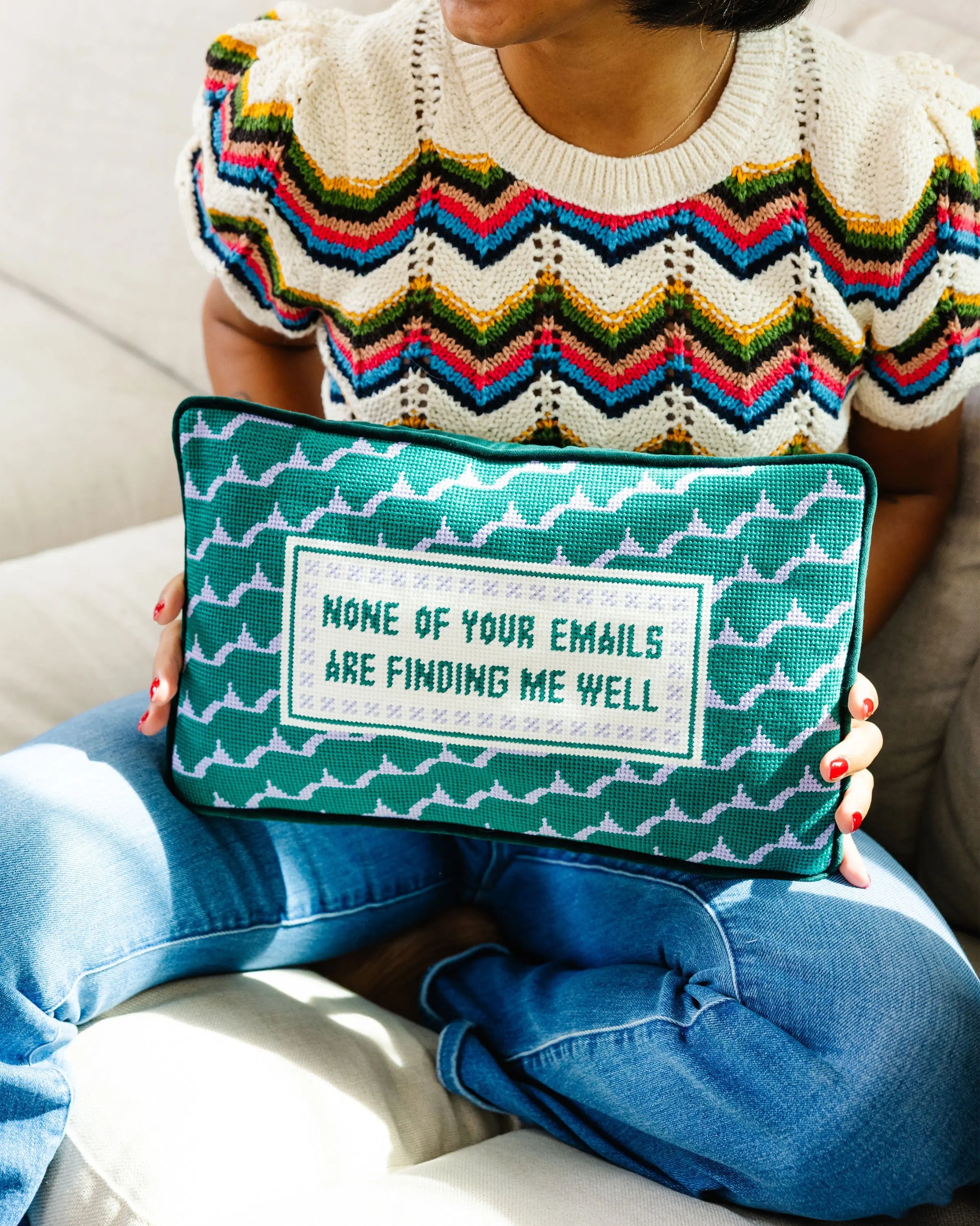 Person holding a green pillow with text, wearing a colorful sweater and blue jeans.