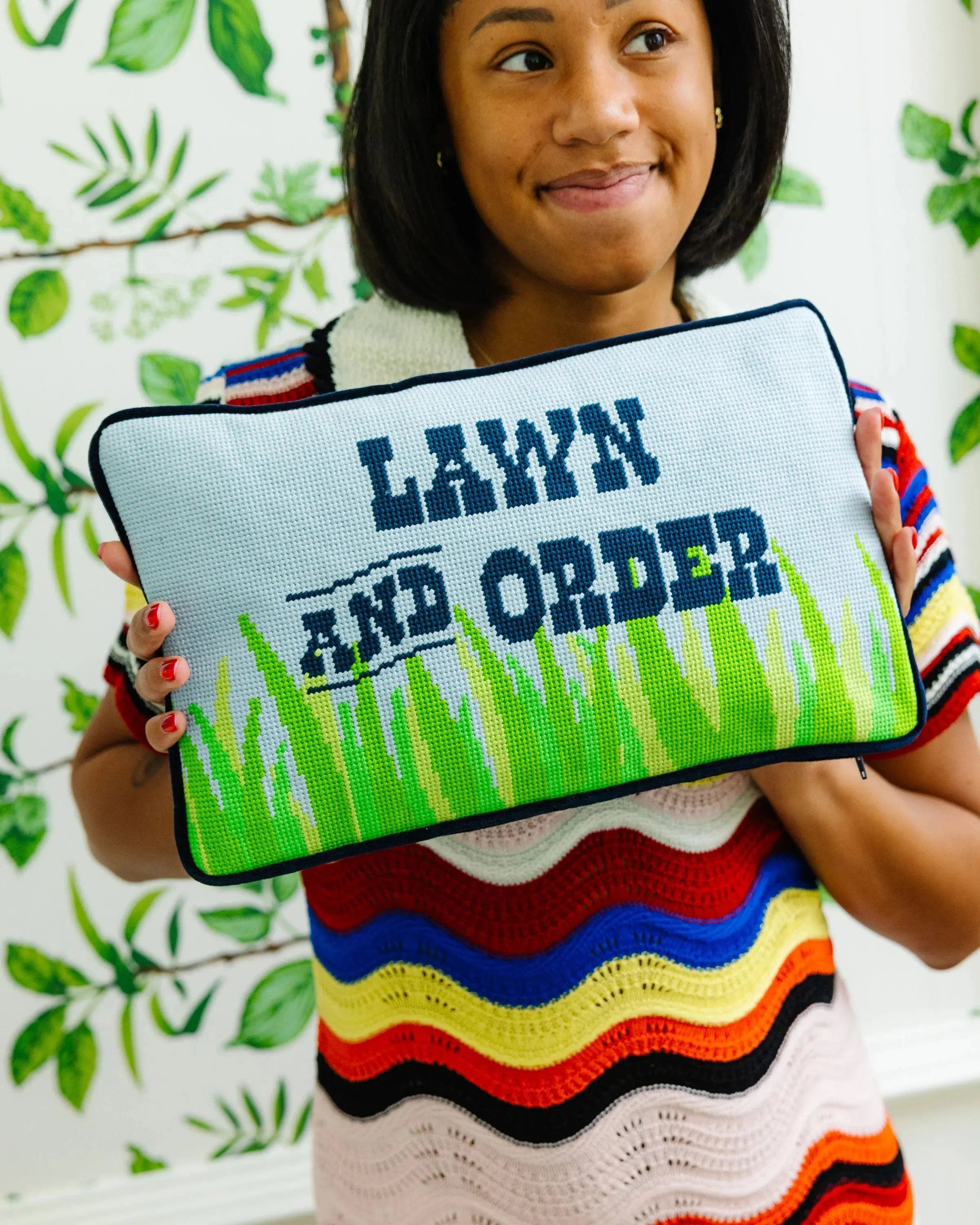 Person holding a cross-stitched pouch with 'Lawn and Order' text against a leafy background