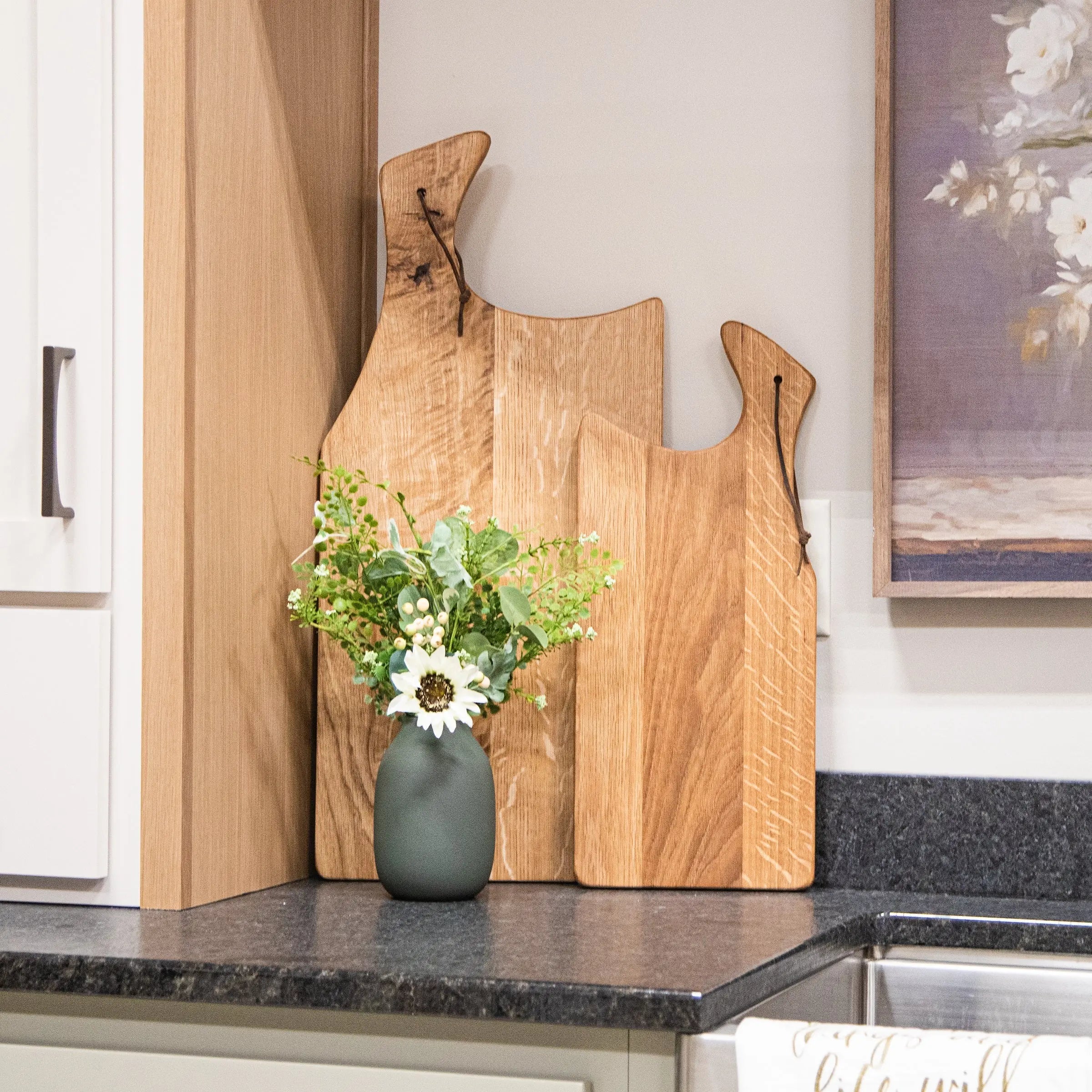 Wooden cutting boards on a kitchen counter with a vase of flowers and a painting in the background.