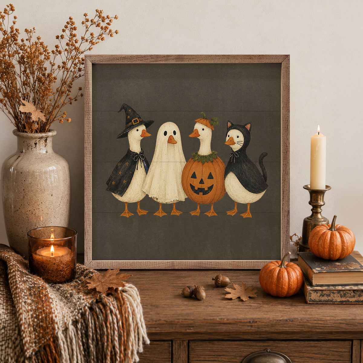 A framed Halloween art print featuring four whimsical goose characters in costumes (witch, ghost, pumpkin, and cat) on a dark gray background, styled on a wooden console table with a candle, pumpkins, acorns, and a vase of dried branches.
