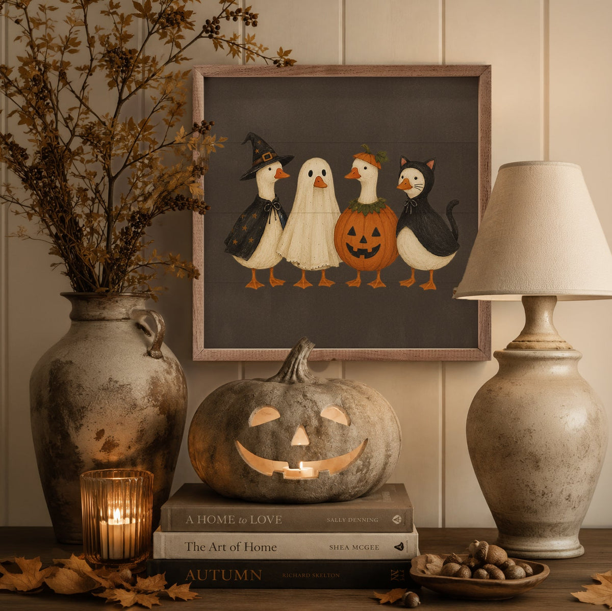A framed art print featuring four cute cartoon pumpkins and ghosts in Halloween costumes, styled in a cozy home decor setting on a wooden table with a carved pumpkin, books, a lamp, and a vase of dried branches.