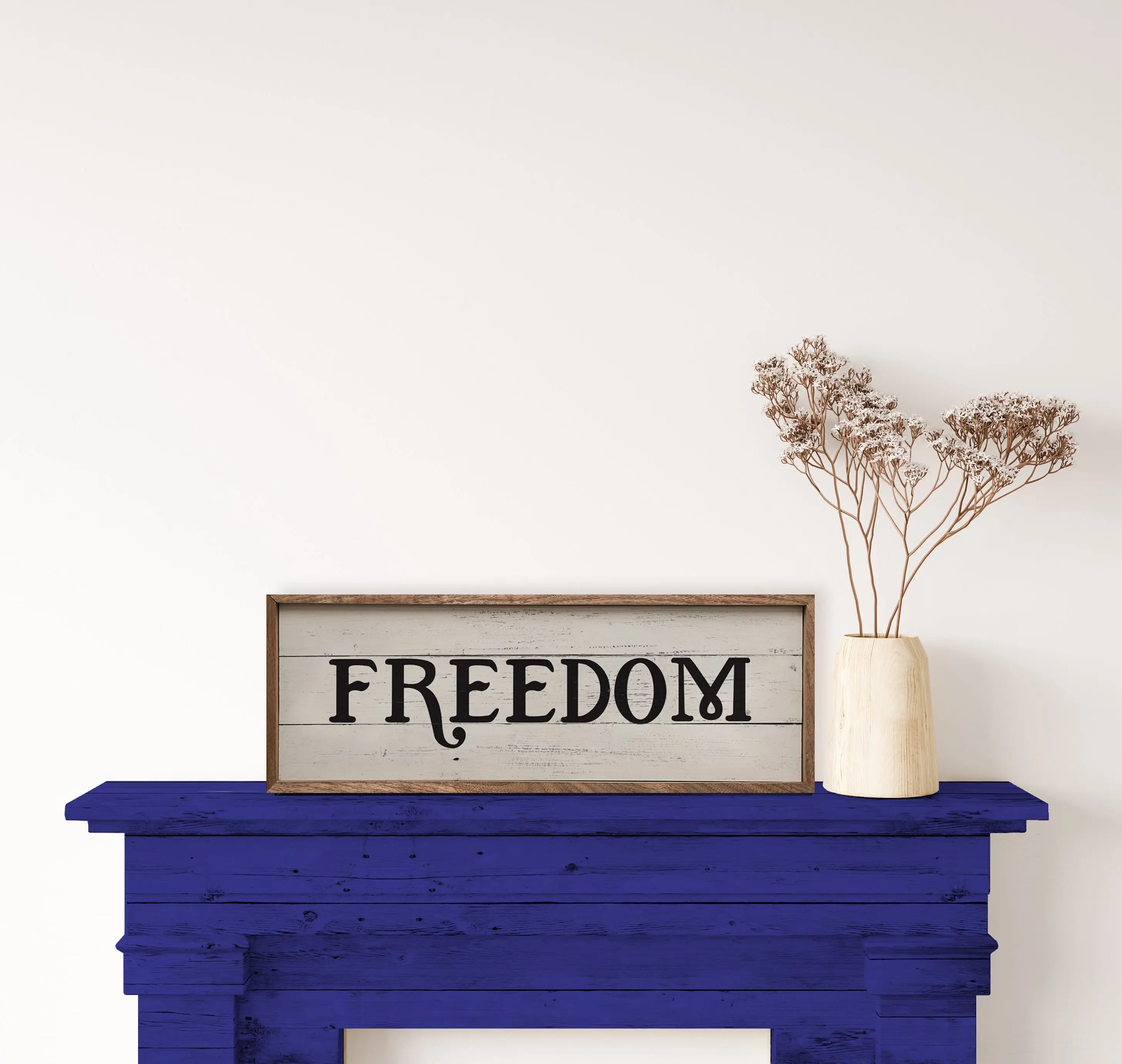Wooden sign with 'FREEDOM' on a blue fireplace mantel with a vase of dried flowers.