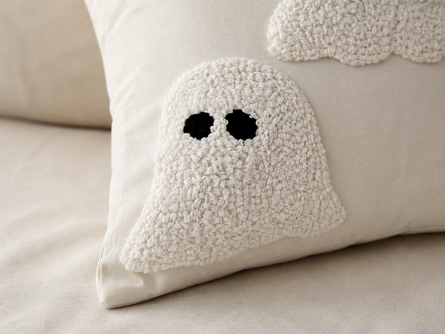 A close-up of a textured cream bouclé throw pillow featuring a ghost applique with black embroidered eyes, resting on a matching beige fabric surface.