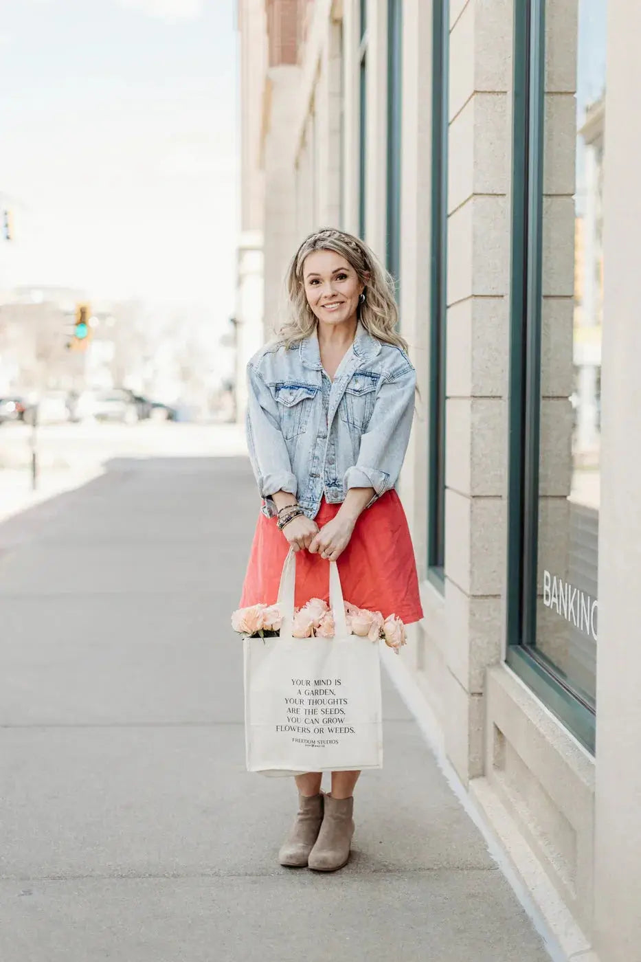 Woman holding tote bag with inspirational quote and pink roses, wearing denim jacket and red skirt on city sidewalk