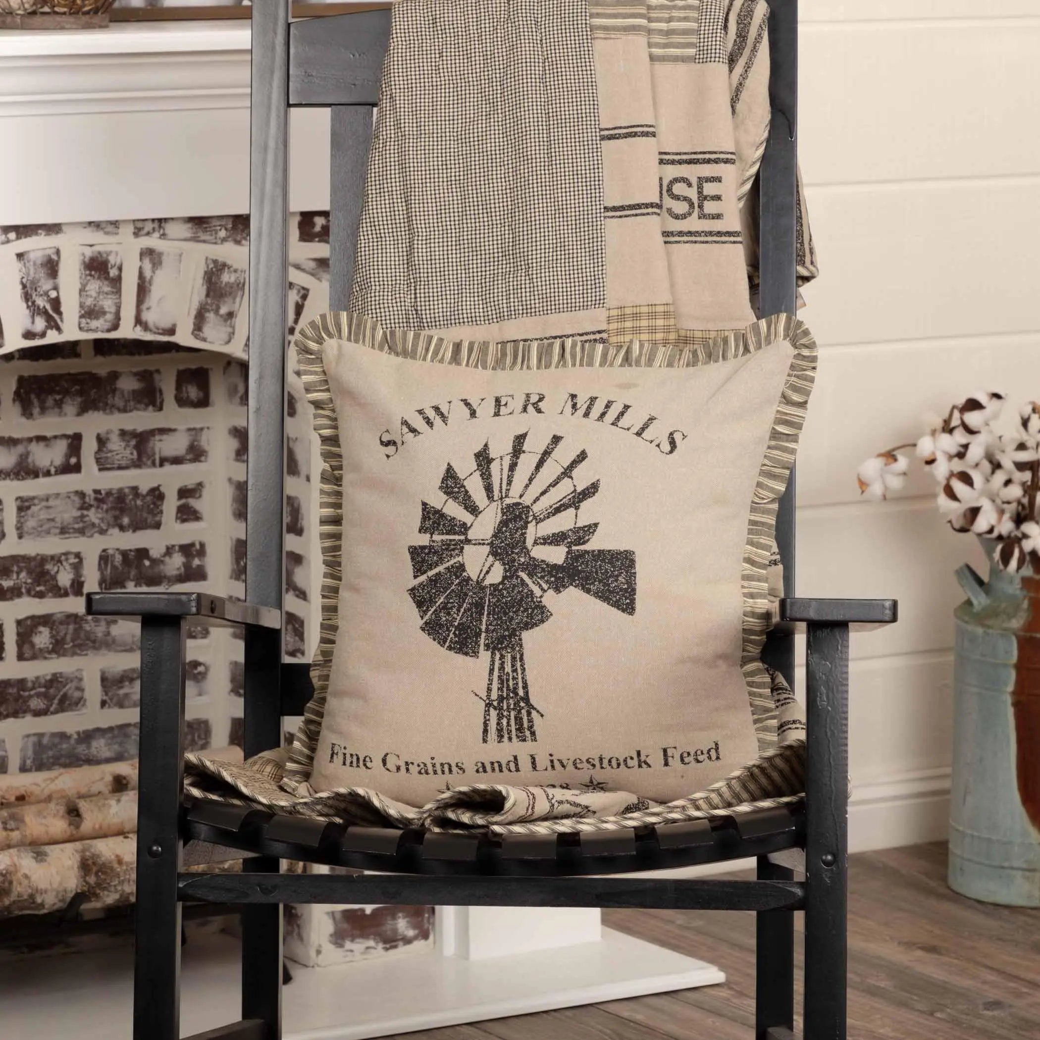 Farmhouse pillow with windmill design and 'Sawyer Mills' text, styled on black chair near fireplace