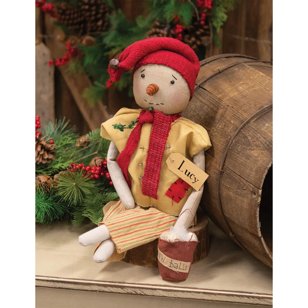A rustic Christmas snowman figurine named 'Lucy' wearing a red hat and scarf, yellow shirt, and striped pants, sitting next to a wooden barrel with pine decorations.