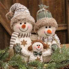 Three plush snowman figurines posed together in front of a rustic wooden backdrop, with evergreen branches at the bottom. The snowmen wear knit hats, striped scarves, and embroidered winter clothing, with one larger snowman, one smaller snowman, and a central snowman figure.