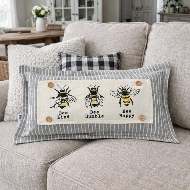 A rectangular lumbar throw pillow with a gray and white striped border and a central cream patch featuring three embroidered bees with the words 'Bee Kind', 'Bee Humble', and 'Bee Happy'. The pillow is styled on a light beige fabric sofa in a cozy, rustic living room setting.