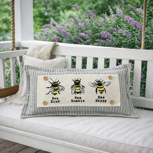 A rectangular decorative throw pillow with a gray and white striped border and a central cream panel featuring three illustrated bees and the words 'Bee Kind,' 'Bee Humble,' and 'Bee Happy.' The pillow is displayed on a white porch swing with cushions, and blurred greenery and purple flowers appear in the background.