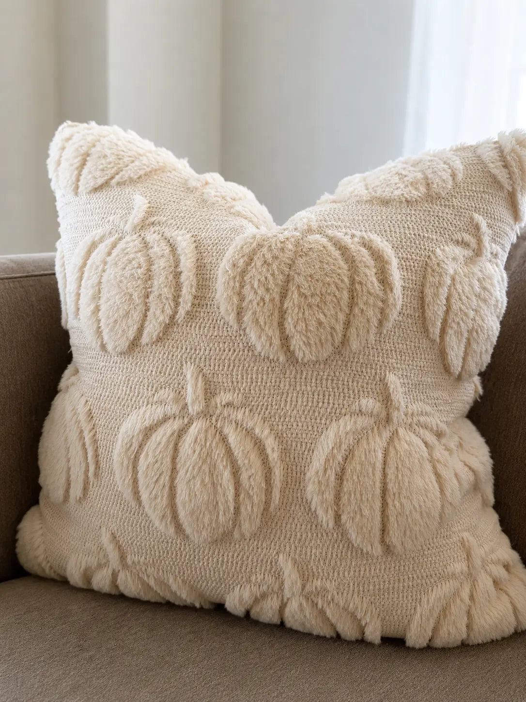 A square cream throw pillow with a textured, fuzzy knit design featuring raised pumpkins and leaves, resting on a brown sofa.