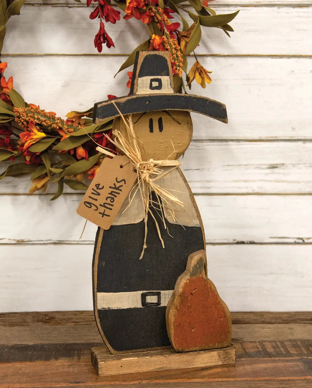 A rustic wooden Thanksgiving-themed decorative figure with a black hat and coat, tan face and hands, a small orange pumpkin at its side, and a twine tag that reads 'give thanks,' displayed against a white wood plank background with a floral wreath.