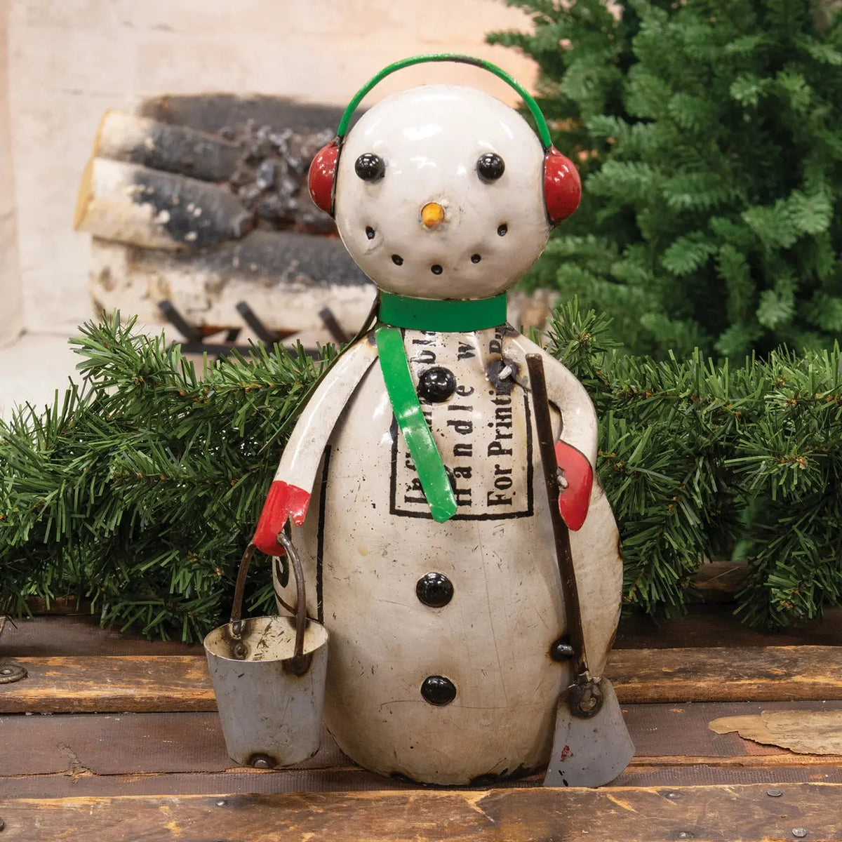 A rustic painted snowman holiday decoration with red earmuffs, a green scarf, and a black-and-white print on its chest, standing on a wooden surface with evergreen branches and logs in the background.