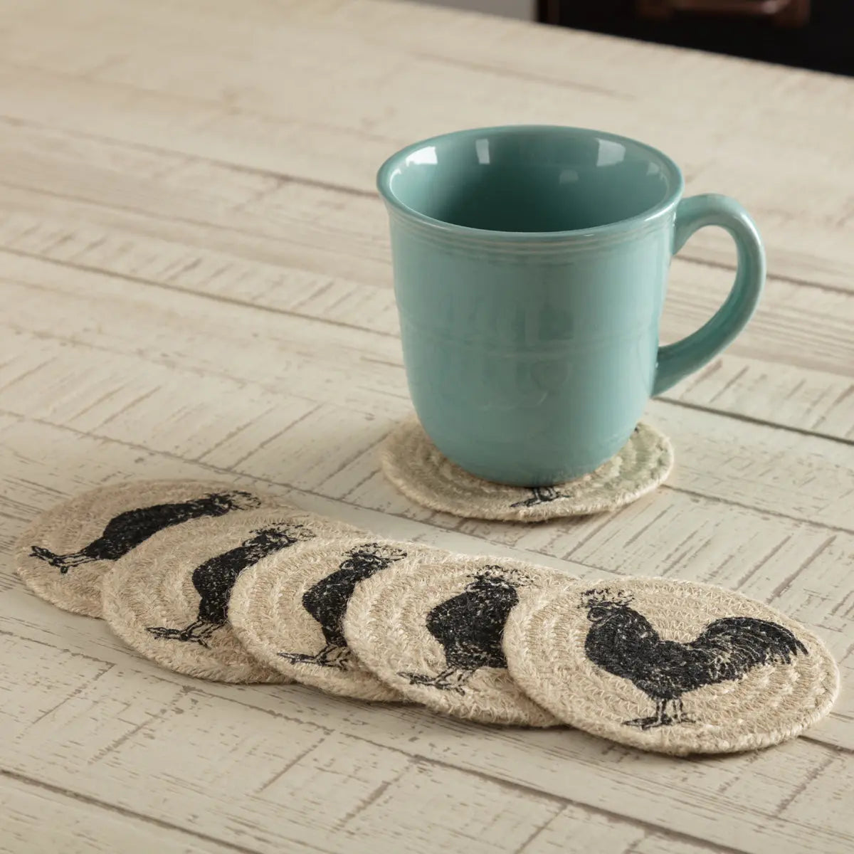 A set of oval woven coasters with black rooster silhouettes, shown on a whitewashed wooden table with a mint green ceramic mug resting on one coaster.
