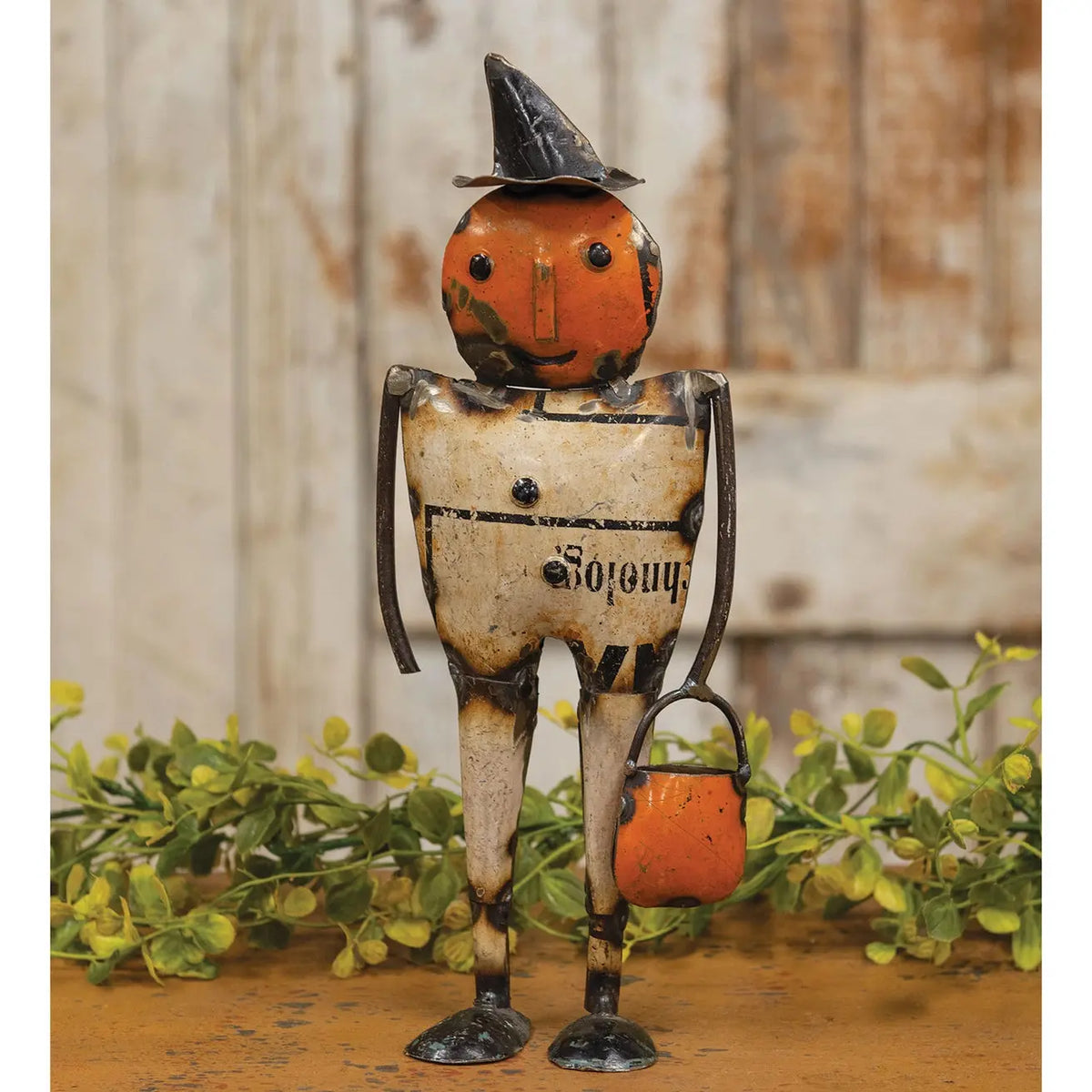 A rustic metal tin figurine of a walking jack-o'-lantern lantern with a black witch hat, holding an orange lantern, set against a weathered wood and green foliage background.