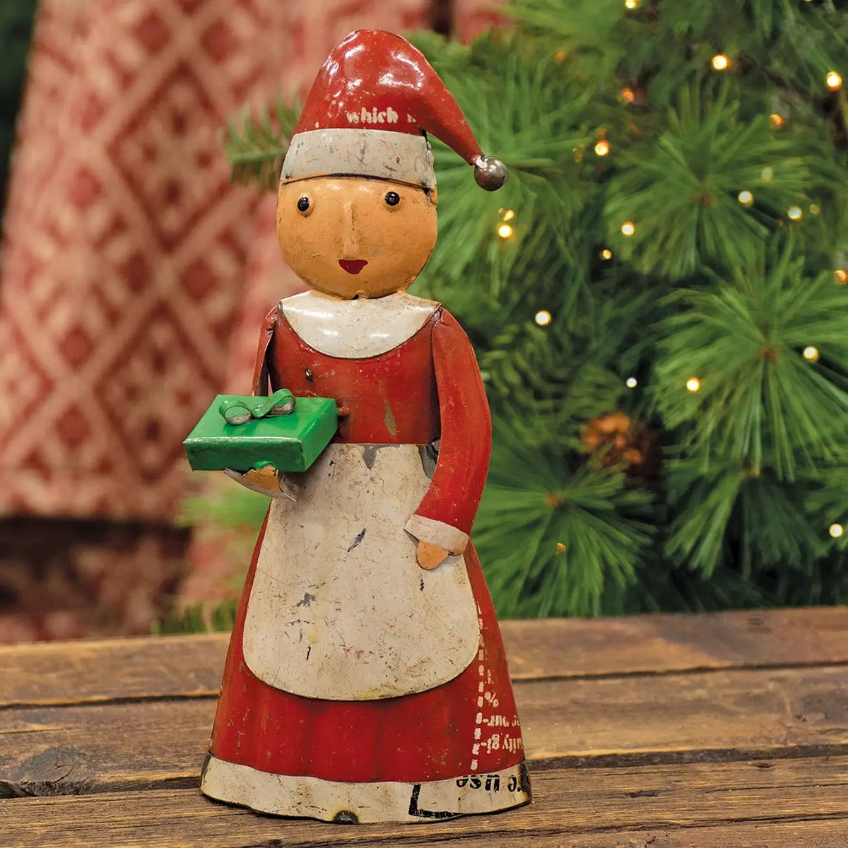 A vintage hand-painted tin Christmas figurine of a woman in a red dress and white apron holding a green gift box, set on a wooden surface with a blurred Christmas tree in the background.