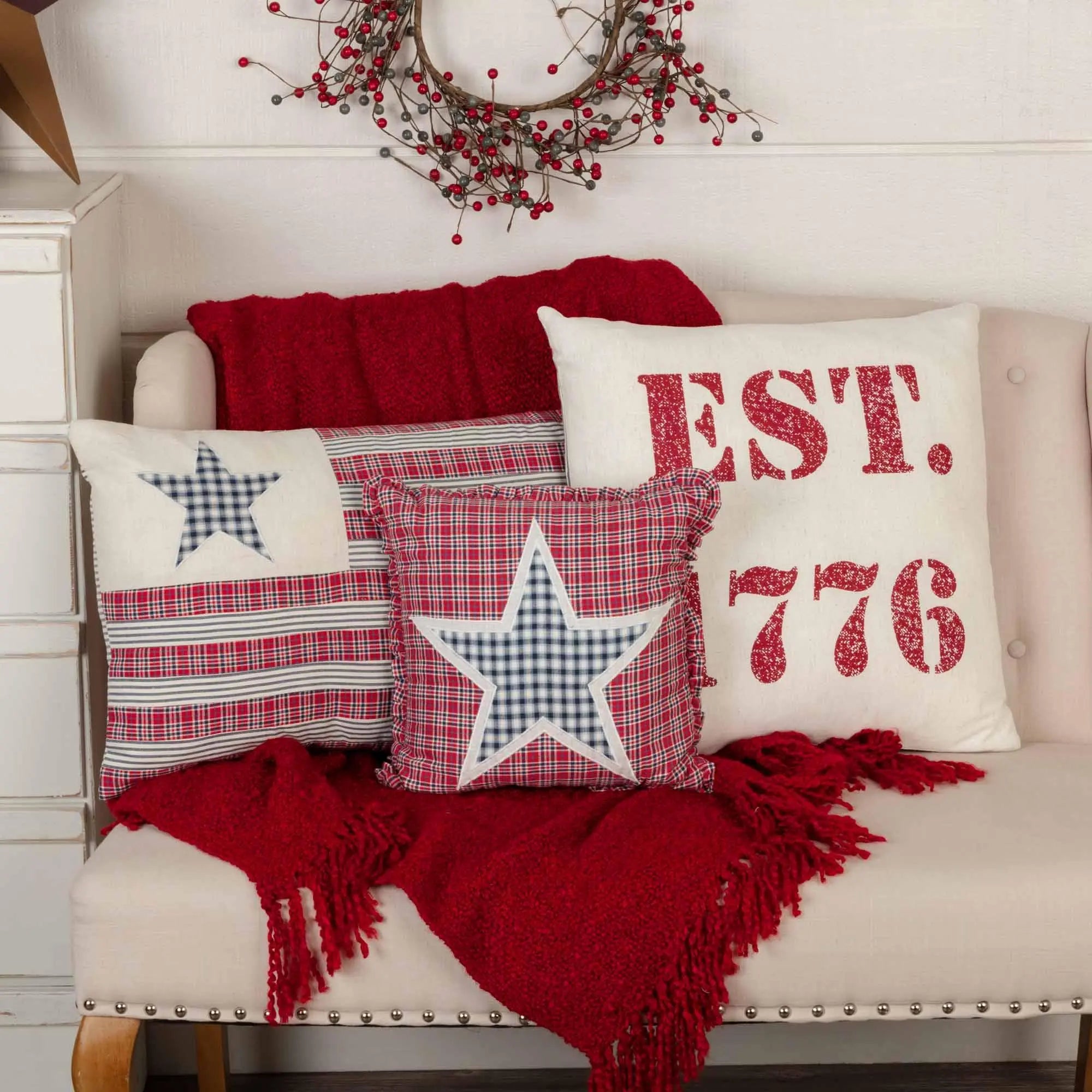 Patriotic farmhouse throw pillows with stars, plaid, and 'EST. 1776' on cozy red blanket, rustic berry wreath above