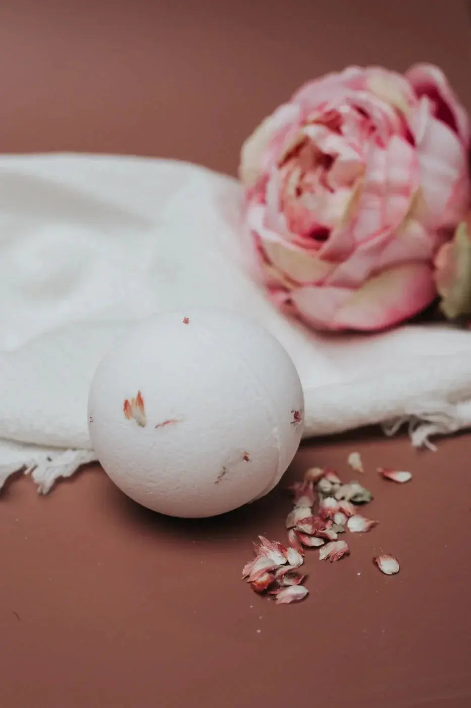White bath bomb with dried rose petals, soft towel, and pink peony for cozy self-care