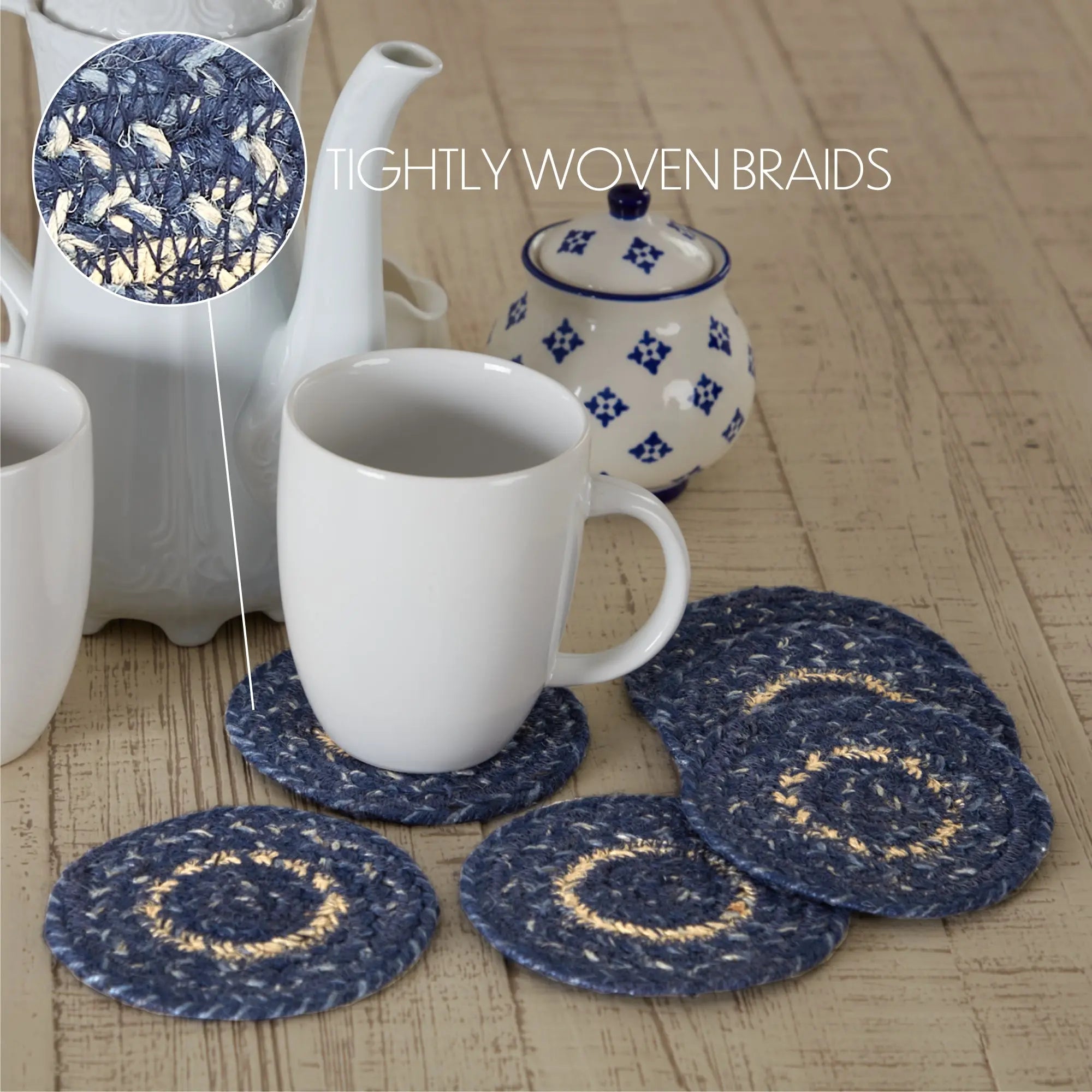 A set of round braided coasters in navy blue and beige woven fabric, shown on a light wood surface with white mugs and a patterned teapot in the background. A zoomed-in inset at the top left highlights the tightly woven braided texture.
