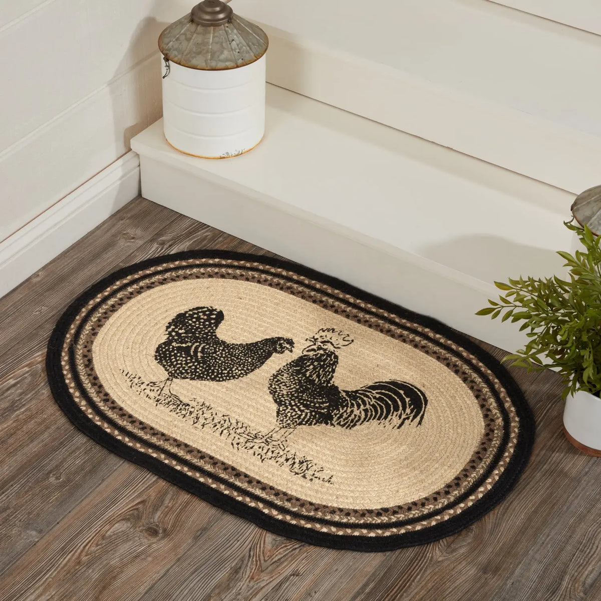 An oval braided jute doormat with a black rooster and chicken design, styled on a rustic wood floor in front of a white step.