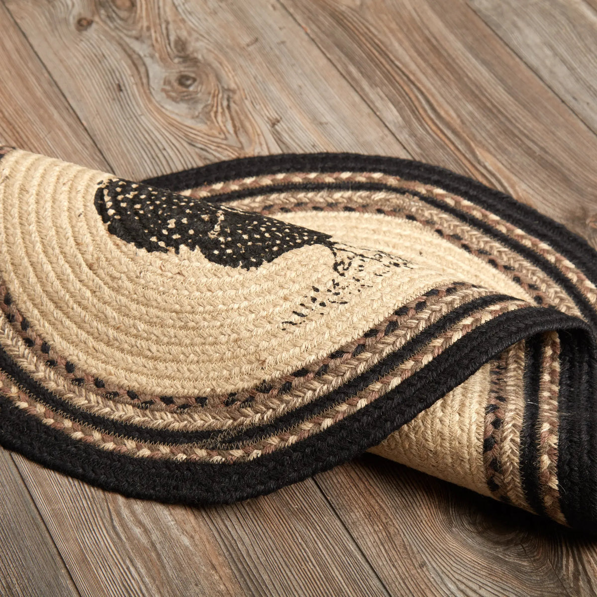 A braided round area rug with a natural tan jute base, black braided borders, and a black woven bird motif, shown partially rolled on a rustic wood plank floor.
