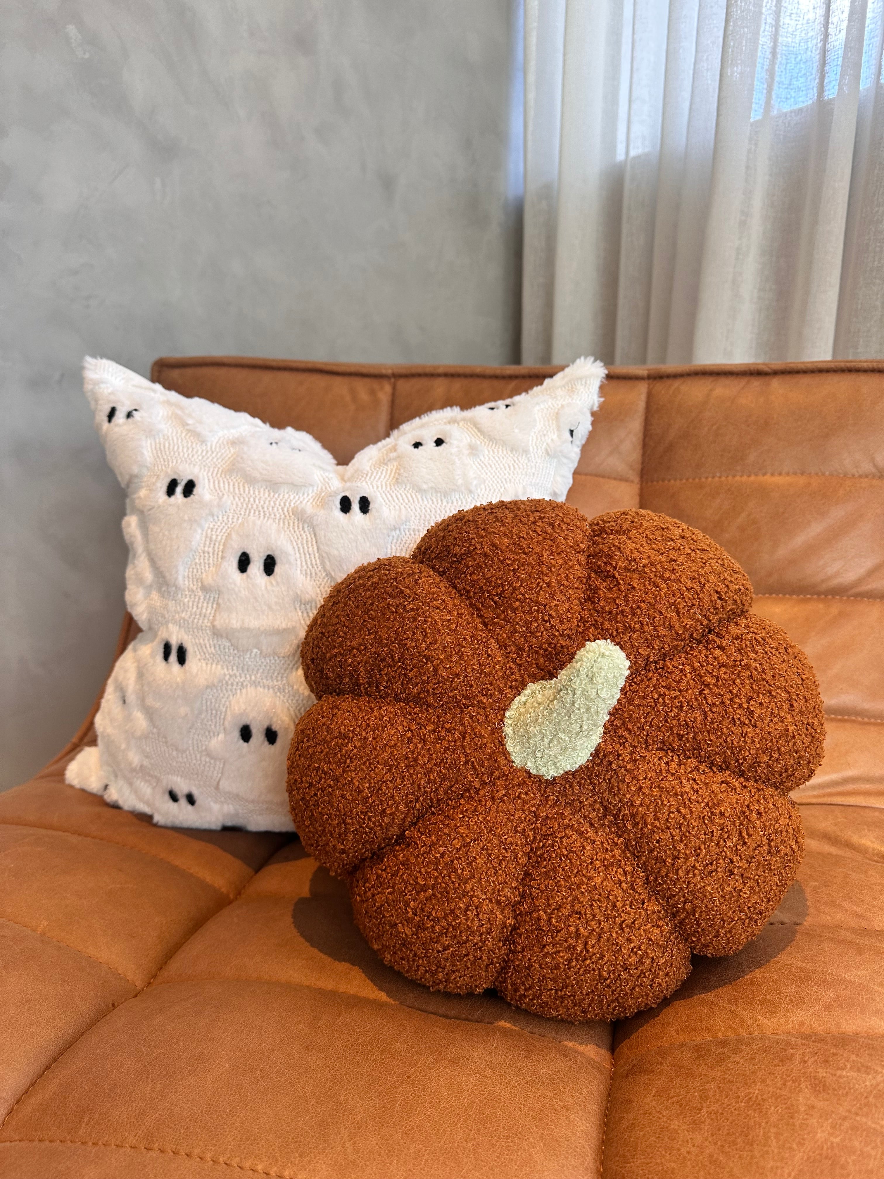 Brown textured pumpkin pillow with a white heart on a brown couch, next to white ghost pillows.