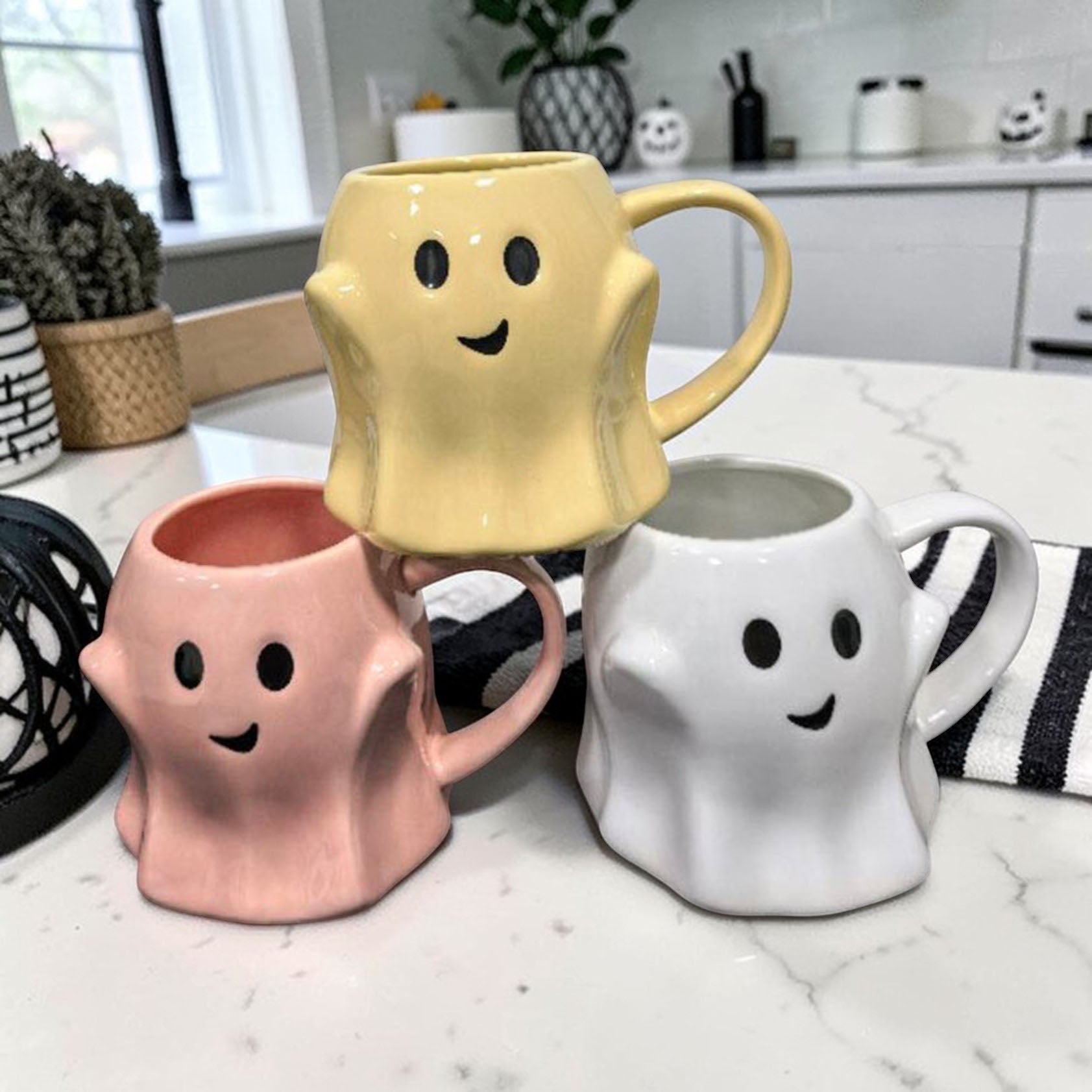 Three ghost-shaped mugs in yellow, pink, and white on a kitchen counter.