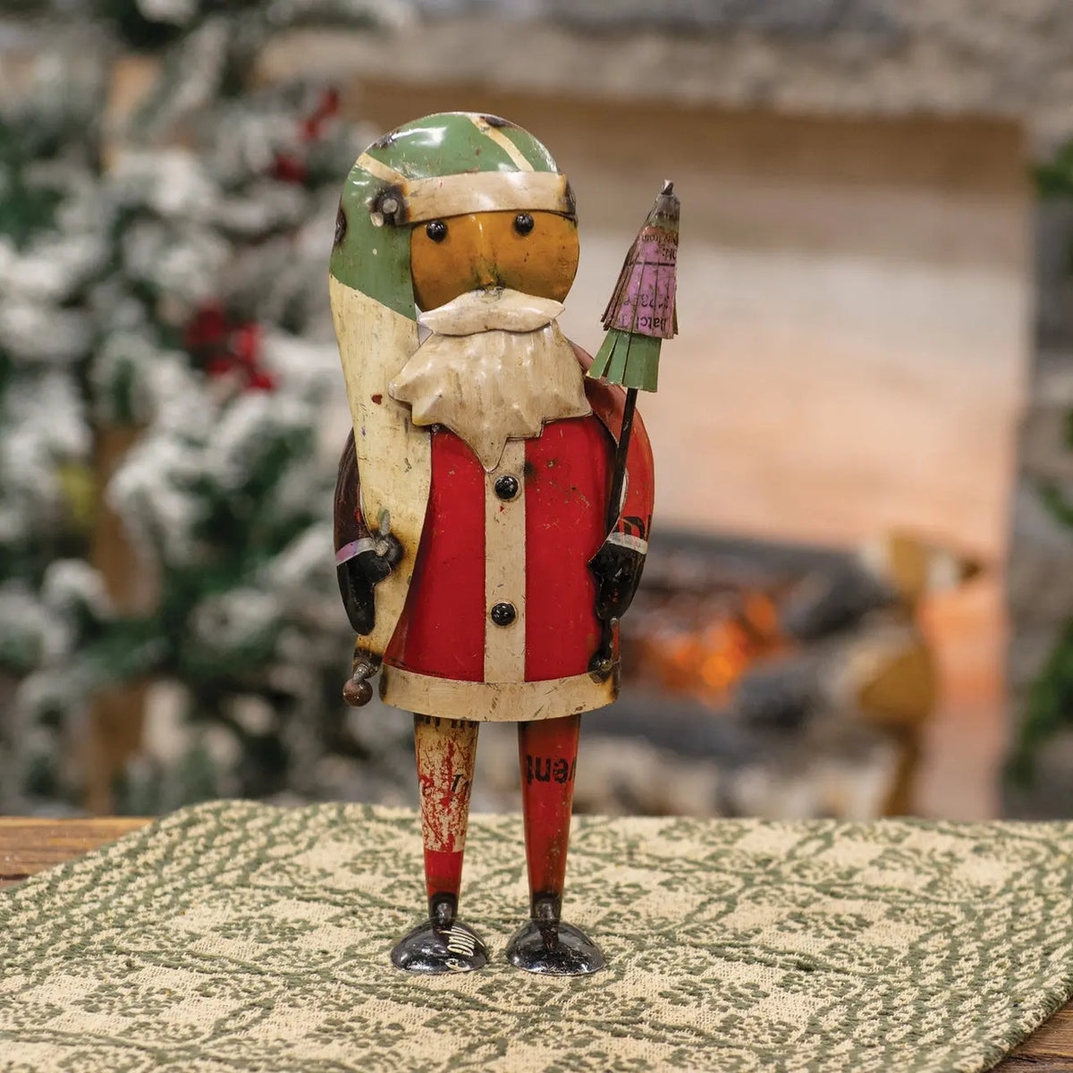 A vintage wooden Santa Claus nutcracker figurine standing on a patterned table runner, with a blurred fireplace and holiday tree in the background.