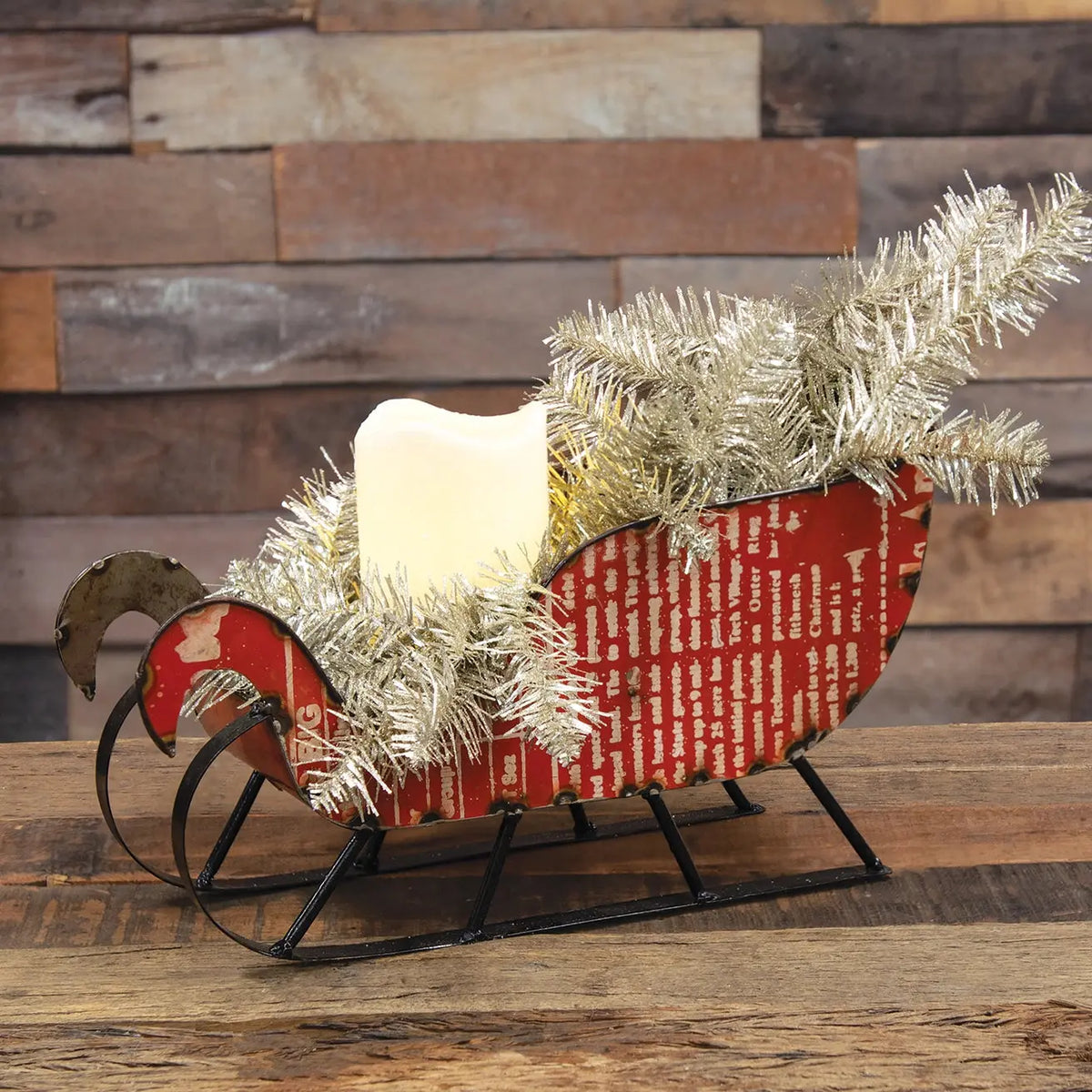 A festive Christmas sleigh-shaped candle holder with a red patterned body, black metal runners, white tinsel garland, and a white candle, displayed on a wooden surface against a rustic wood plank wall.