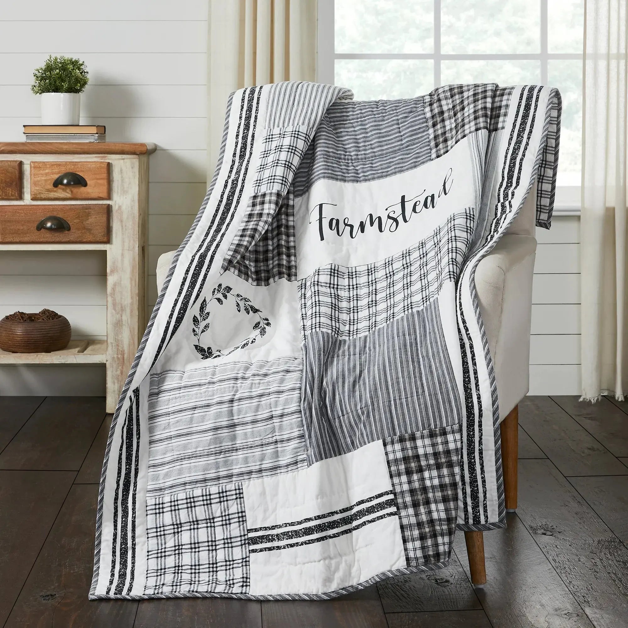 Patchwork blanket with 'Farmstead' text draped over a chair in a room with wooden floor and white walls.