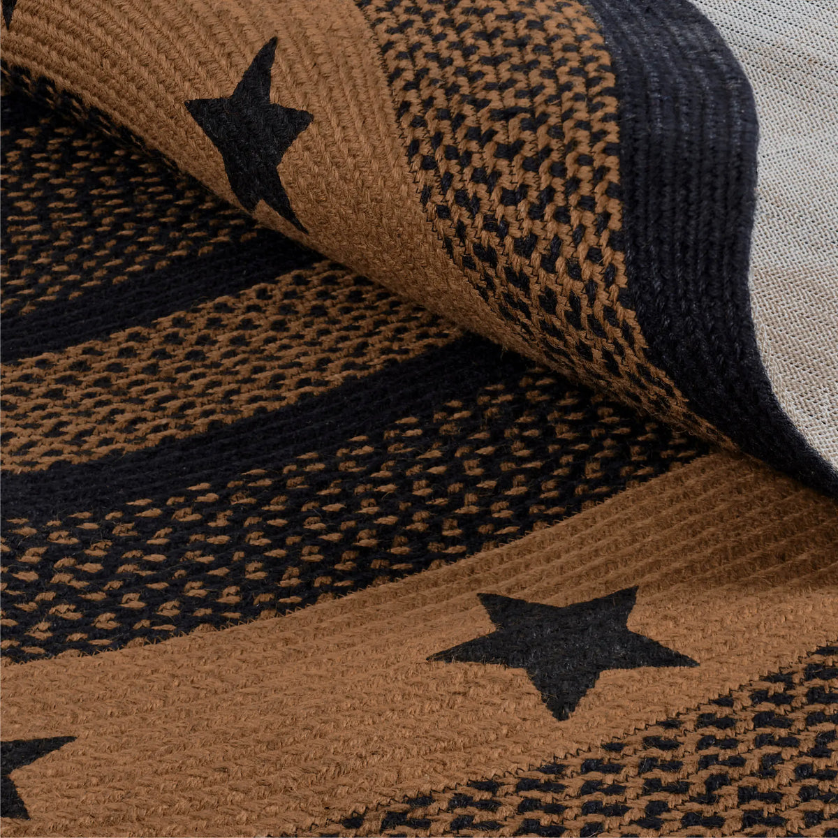 A close-up of a woven textile with a tan and black pattern featuring textured stripes and black star motifs, shown with one corner folded over to reveal the weave.
