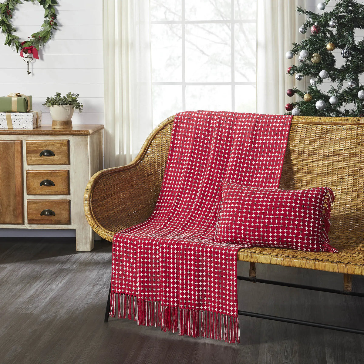 A red and white patterned knit throw blanket with fringe is draped over a wicker sofa, with a matching throw pillow. The scene is set in a decorated living room with a Christmas tree, a wooden cabinet, and a wreath.