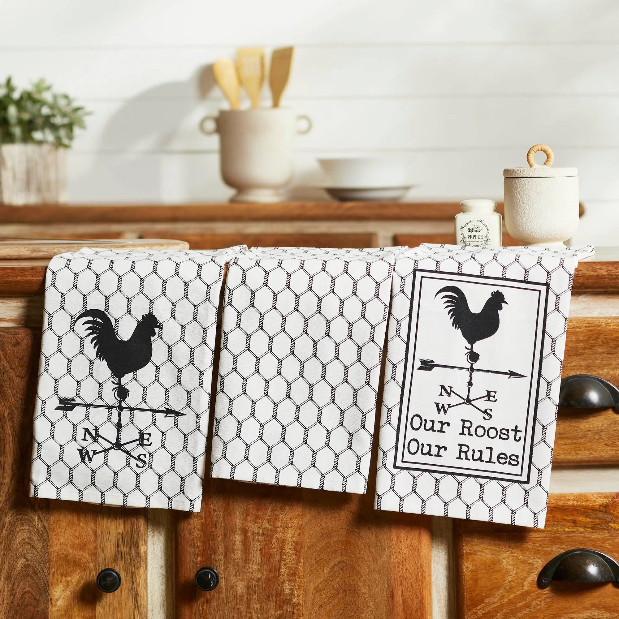 Three kitchen towels with rooster designs on a wooden cabinet.