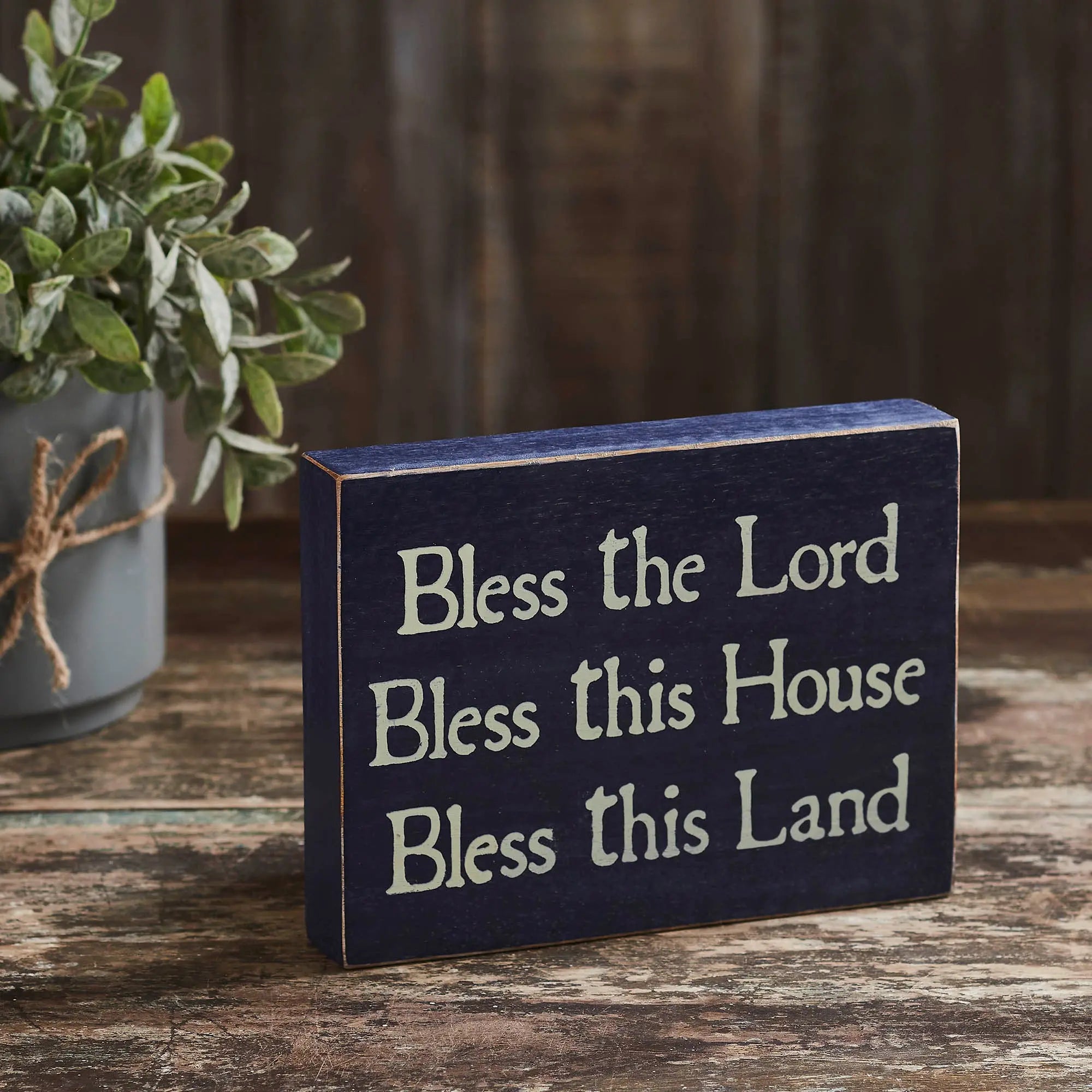 Decorative sign with text 'Bless the Lord Bless this House Bless this Land' on a wooden surface.
