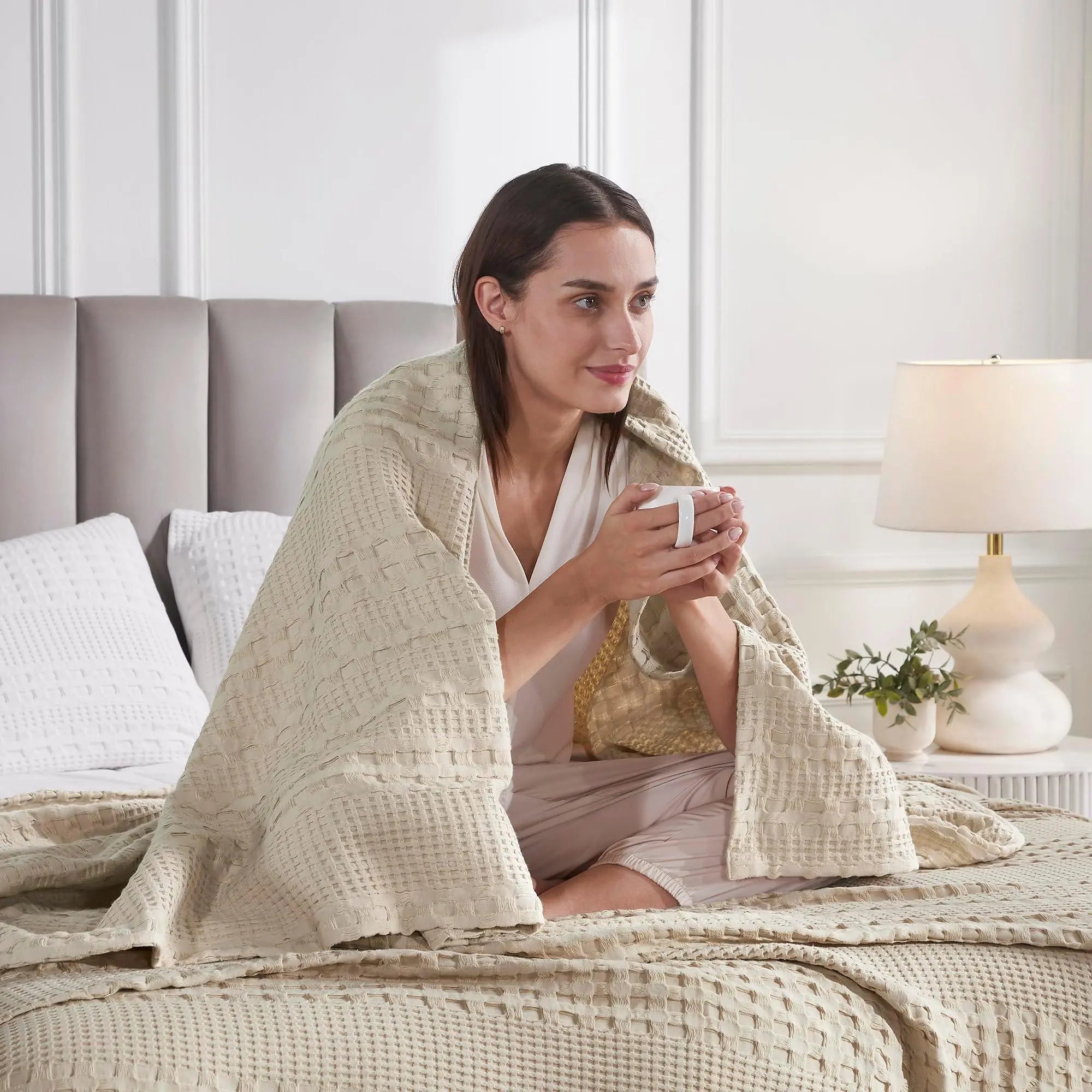 Woman wrapped in cozy beige waffle knit blanket on bed, holding mug, farmhouse bedroom decor