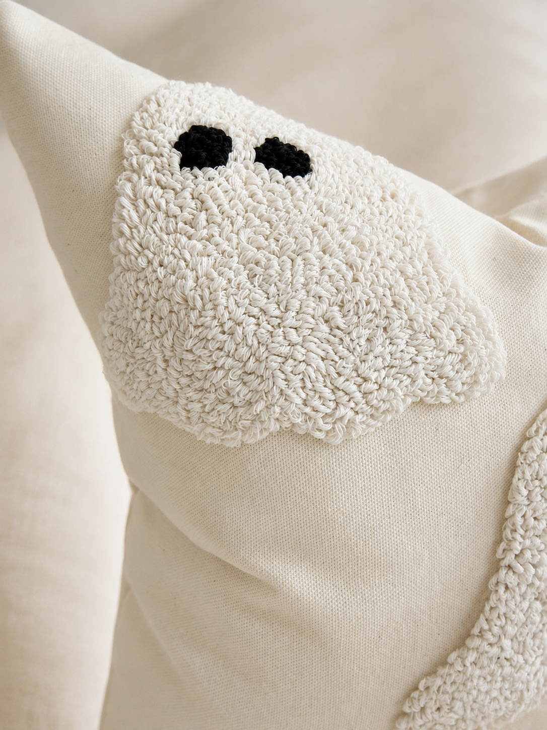 Close-up of a cream-colored textured throw pillow with a ghost face design, featuring black embroidered eyes and a chunky terry cloth patch.