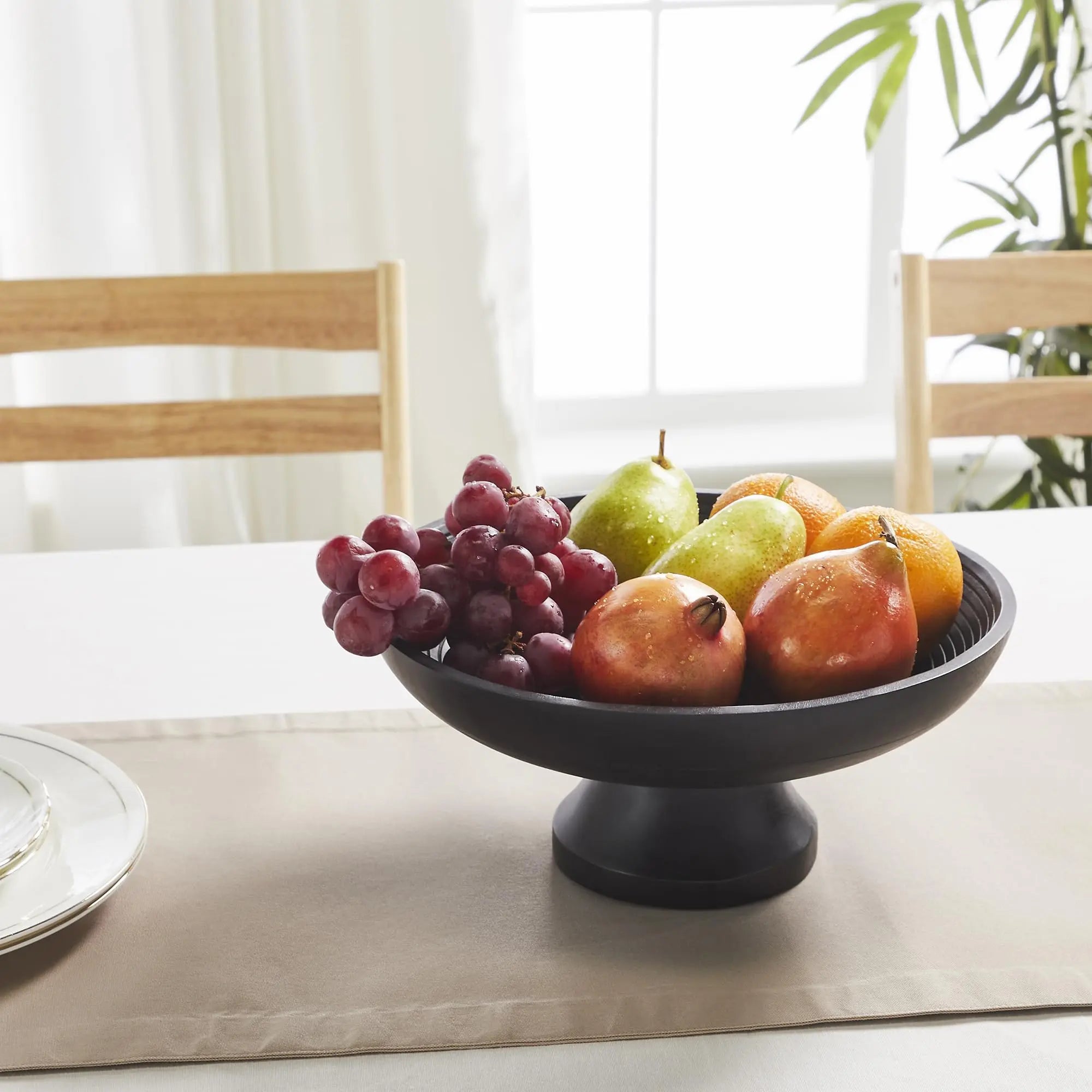 Fruit bowl with grapes, pears, and apples on a dining table with chairs and a plant in the background.