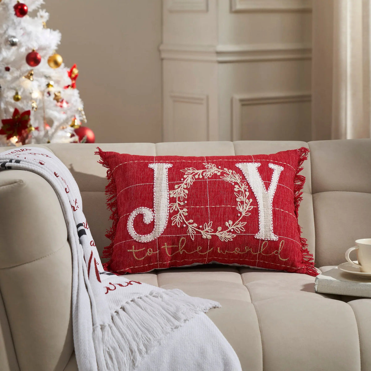 A red rectangular throw pillow with 'JOY to the world' embroidered in white and gold, styled on a beige sofa next to a white Christmas tree and a white throw blanket.