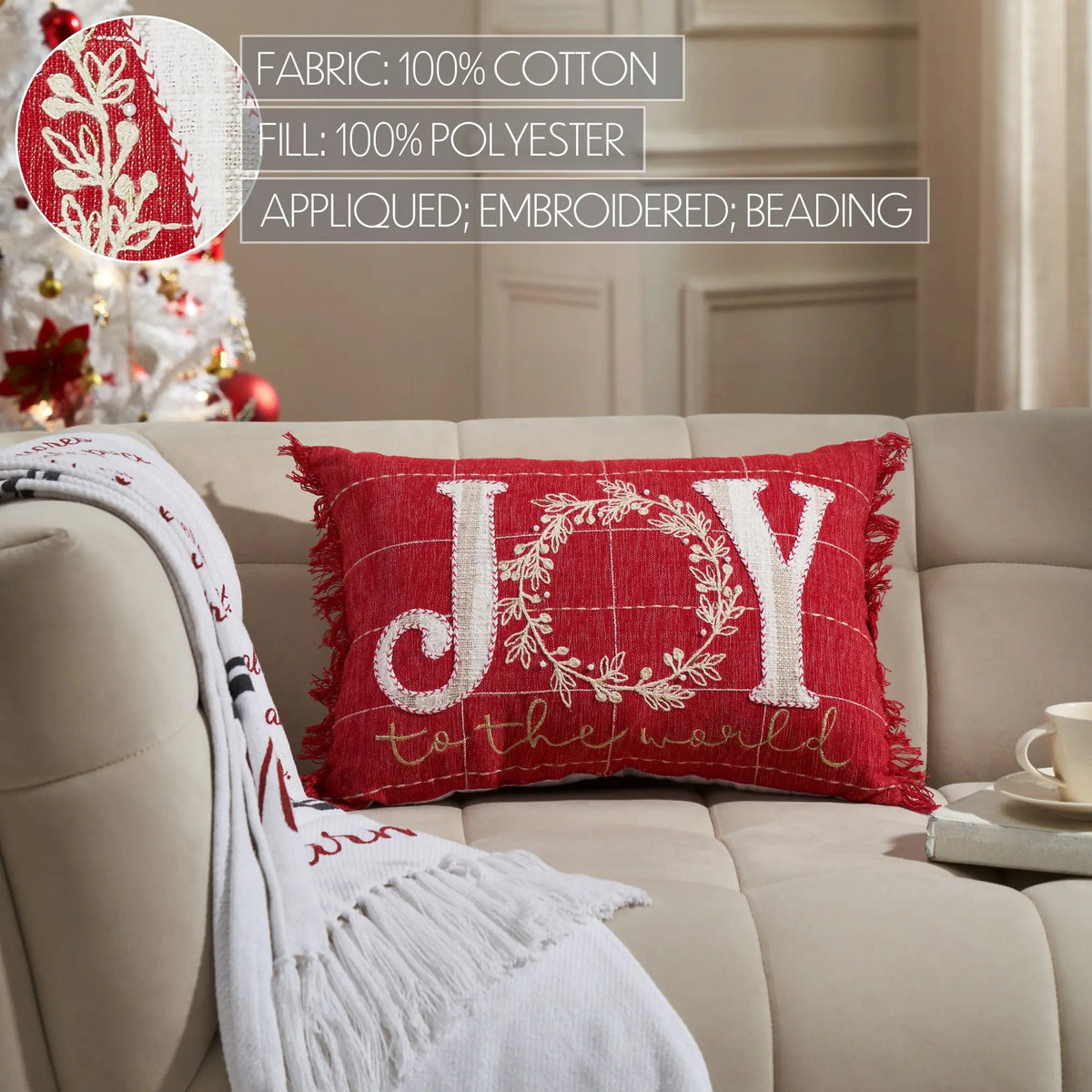 Red 'JOY' pillow with white embroidery on a beige couch, Christmas tree in the background.