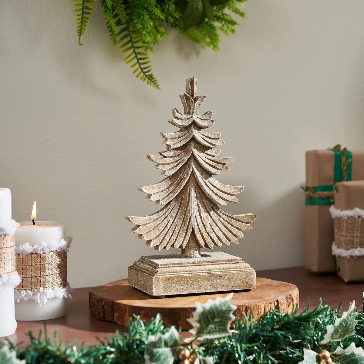 A small carved wooden Christmas tree figurine with a distressed beige finish, displayed on a round wood slice coaster. The scene is decorated with holiday greenery, a lit white candle, and wrapped gift boxes in the background.