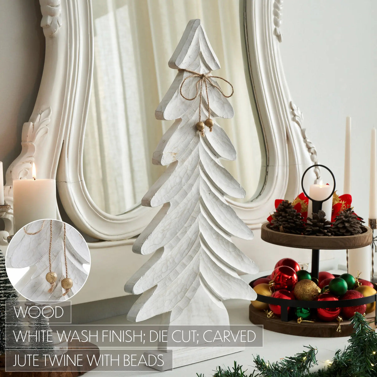 A white-washed wooden Christmas tree ornament with carved layered branches, a jute twine bow, and small bead accents, displayed on a tabletop beside candles, a mirror, and holiday decorations.