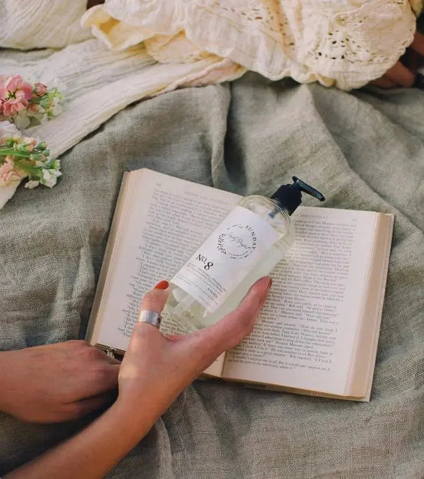 Cozy farmhouse bath oil bottle held over open book with soft linens and flowers, perfect for relaxation and self-care